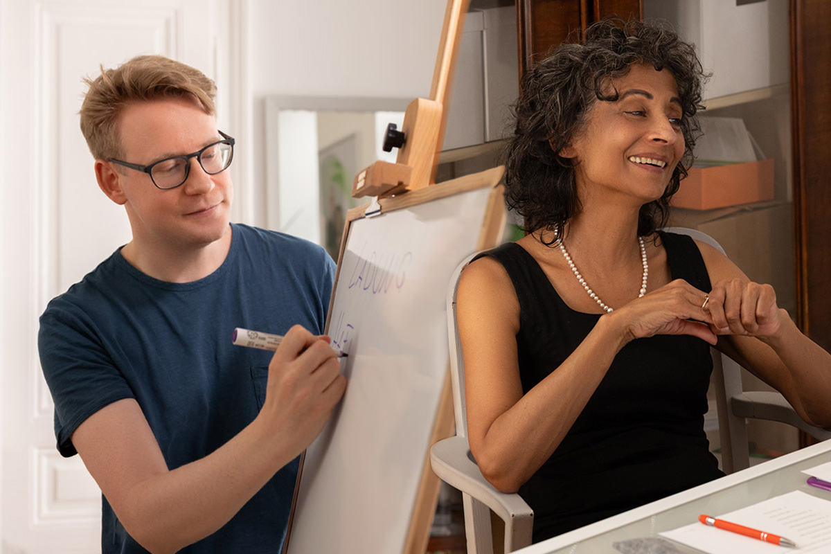 Michael En schreibt auf einem Whiteboard; Michèle Cooke sitzt und lächelt.